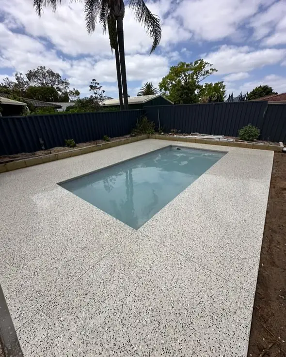 Full backyard pool surround in white exposed aggregate with palms and a Colorbond fence behind