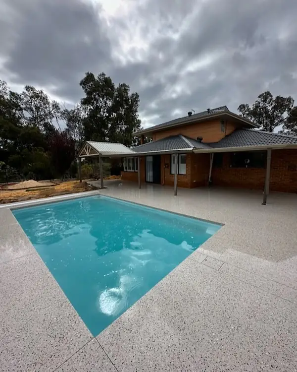 Exposed aggregate pool surround wrapping around a blue pool beside a brick two storey house under a grey sky